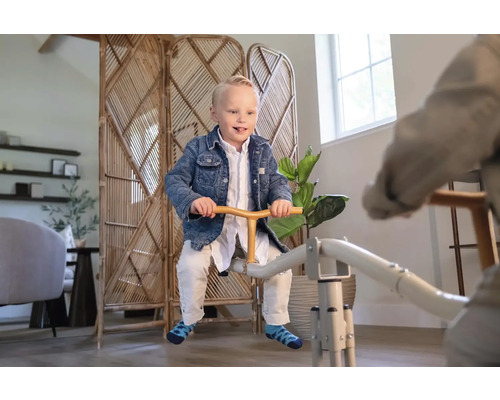 Ein Kind sitzt auf einer Metallwippe in einem hellen, modern eingerichteten Raum mit einem Rattan-Paravent und Zimmerpflanzen.