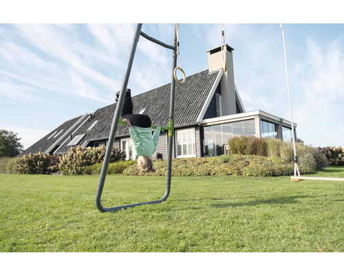 Spielgerät aus Metall im Garten mit Turnreck, Gymnastikringen und Schaukel, Kind turnt kopfüber an der Stange vor einem Haus mit Rasenfläche.
