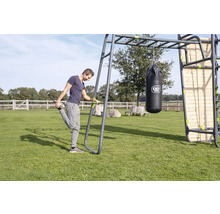 Ein Mann dehnt sich an einer EXIT Toys Outdoor-Fitnessstation mit Hangelgriff, Boxsack und Holzkletterwand auf einer Rasenfläche.