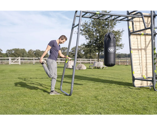 Ein Mann dehnt sich an einer EXIT Toys Outdoor-Fitnessstation mit Hangelgriff, Boxsack und Holzkletterwand auf einer Rasenfläche.