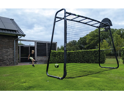 Ein Kind schießt einen Fußball in ein schwarzes Multifunktions-Fußballtor mit Basketballkorb auf einem grünen Rasen im Garten.