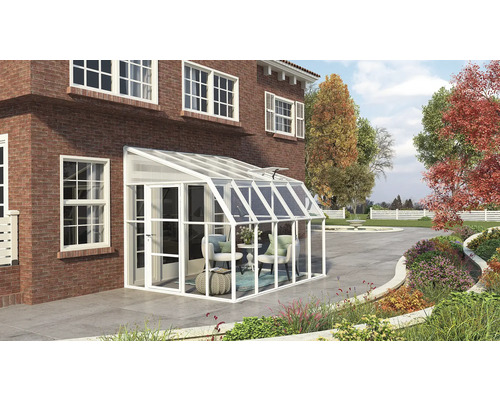 Weißes Anlehngewächshaus an einer roten Backsteinwand mit Gartenmöbeln und bepflanztem Außenbereich.