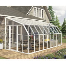 Weißes Anlehngewächshaus aus Aluminium an einer Backsteinwand auf einer Holzterrasse, eingerichtet mit Tisch, Stühlen und einem Sofa.