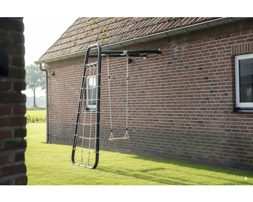 Wandmontiertes Spielgerät mit schwarzem Metallrahmen, einer Holzschaukel und einem Kletternetz an einer Ziegelwand im Garten.