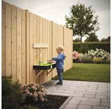 Wandmontierte Matschküche aus Holz an einem Gartenzaun in einem hellen Garten mit einem spielenden Kind auf einer Terrasse.