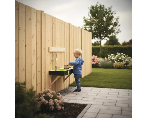 Wandmontierte Matschküche aus Holz an einem Gartenzaun in einem hellen Garten mit einem spielenden Kind auf einer Terrasse.