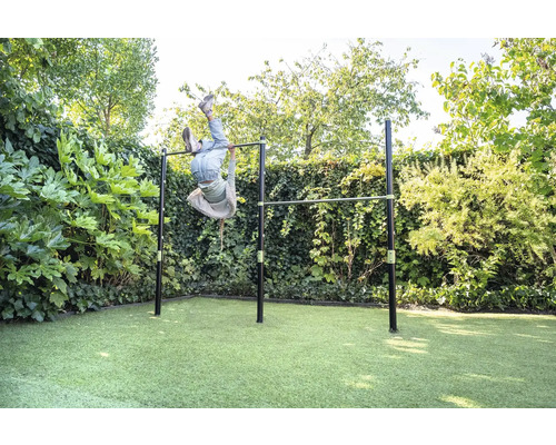 Doppel-Reckstange aus Metall im Garten, ein Kind turnt kopfüber an der Stange, umgeben von Hecken und Rasen, helle und weitläufige Atmosphäre.