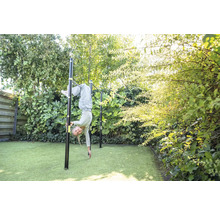 Ein Kind turnt an einer schwarzen Reckstange aus Metall in einem hellen, grünen Garten mit Kunstrasen und dichten Sträuchern.