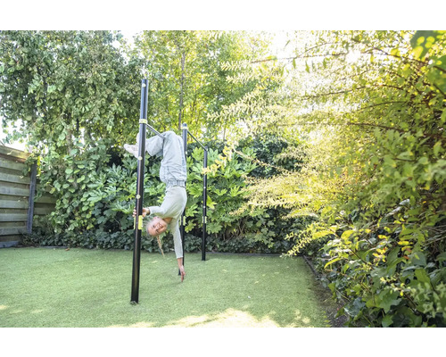 Ein Kind turnt an einer schwarzen Reckstange aus Metall in einem hellen, grünen Garten mit Kunstrasen und dichten Sträuchern.