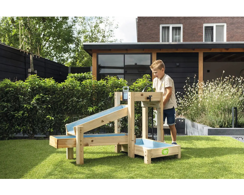 Ein Kind spielt mit einem hölzernen Wasserspielset von EXIT Toys im Garten. Das Set besteht aus mehreren Rinnen und einem Becken. EXIT Toys Logo.