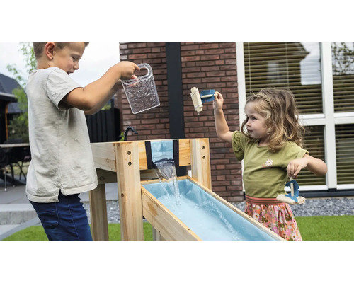 Zwei Kinder spielen an einer Wasserbahn aus Holz im Garten. Ein Junge gießt Wasser aus einem Krug, ein Mädchen spielt mit kleinen Holzbooten.