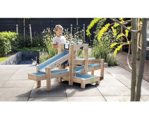 Ein Kind spielt im Garten mit einer mehrstufigen Wasserbahn aus Holz mit blauen Kunststoffrinnen auf einer gepflasterten Terrasse.