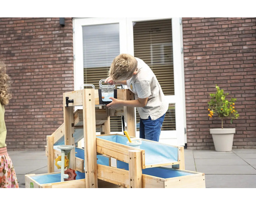 Kind spielt an einer hellen Wasserlandschaft aus Holz mit mehreren Ebenen und blauen Becken auf einer gepflasterten Terrasse.