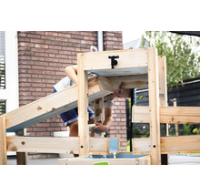 Kind spielt an einer Wasserbahn aus Holz mit Wasserhahn und Rinnen im Garten vor einer Ziegelwand.