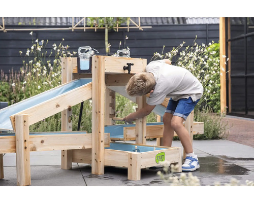 Kind spielt mit einer hölzernen Wasserbahn im Garten. Die Spielanlage umfasst mehrere Ebenen mit Holzrinnen und Wasserbecken für draußen.