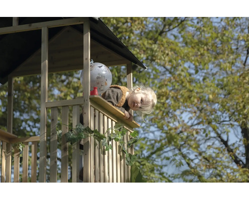 Kleinkind schaut vom Balkon eines hölzernen Spielhauses im Garten.