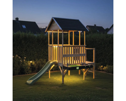 Beleuchtetes Stelzenhaus aus Holz für Kinder mit Rutsche, Dach und integrierter Spielküche im Garten bei Dämmerung.