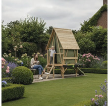 Kinderspielhaus aus Holz im Garten mit Leiter, Balkon und integriertem Spieltisch, umgeben von Blumen und Rasen, mit einem Kind und einer Frau.