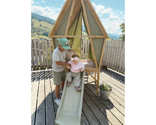 Holz-Spielhaus mit Rutsche von EXIT Toys auf einer Terrasse. Ein Junge hilft einem Kleinkind. Dreieckiger Holzrahmen mit Stoffwänden.