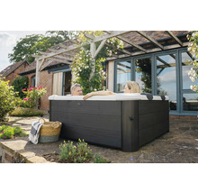 Zwei Personen entspannen in einem quadratischen Außenwhirlpool mit dunkelgrauer Holzverkleidung auf einer Terrasse neben einem Haus mit Pergola.