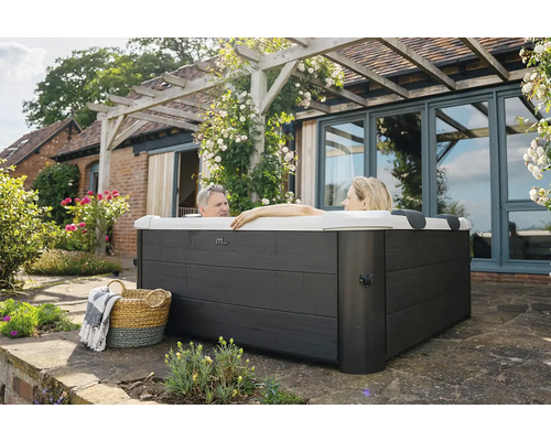 Zwei Personen entspannen in einem quadratischen Außenwhirlpool mit dunkelgrauer Holzverkleidung auf einer Terrasse neben einem Haus mit Pergola.
