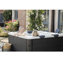Ein Paar entspannt in einem modernen, rechteckigen Outdoor-Whirlpool mit dunkler Holzoptik-Verkleidung in einem Garten mit Blumen.