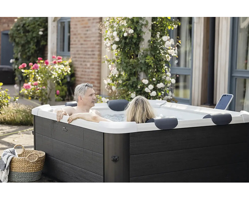 Ein Paar entspannt in einem modernen, rechteckigen Outdoor-Whirlpool mit dunkler Holzoptik-Verkleidung in einem Garten mit Blumen.