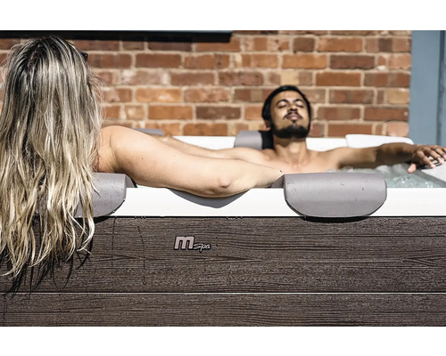 Ein Mann und eine Frau entspannen in einem MSpa Whirlpool mit Außenverkleidung in Holzoptik vor einer Ziegelwand.