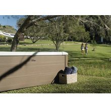 MSpa Whirlpool mit Paneelen in Holzoptik in einem weitläufigen Garten mit Rasenfläche und Bäumen. Ein Korb mit Handtüchern steht daneben.