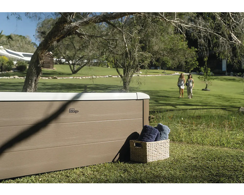 MSpa Whirlpool mit Paneelen in Holzoptik in einem weitläufigen Garten mit Rasenfläche und Bäumen. Ein Korb mit Handtüchern steht daneben.