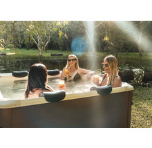 Drei Frauen entspannen in einem rechteckigen Außen-Whirlpool mit Kopfstützen in einem Garten an einem Teich.