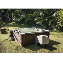 Quadratischer Whirlpool mit Verkleidung in Holzoptik und externer Pumpeneinheit, auf einem Rasen in einem Garten mit Palmen.