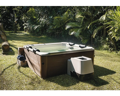 Quadratischer Whirlpool mit Verkleidung in Holzoptik und externer Pumpeneinheit, auf einem Rasen in einem Garten mit Palmen.