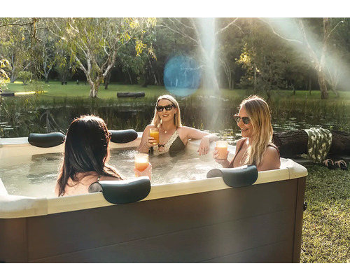 Drei Frauen entspannen in einem Whirlpool im Garten neben einem Teich. Der Whirlpool hat eine dunkle Verkleidung und Kopfstützen.