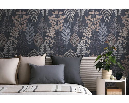 Vliestapete mit botanischem Blättermuster in Dunkelblau und Grau hinter einem Bett mit Kissen und einer Zimmerpflanze auf einem Beistelltisch.