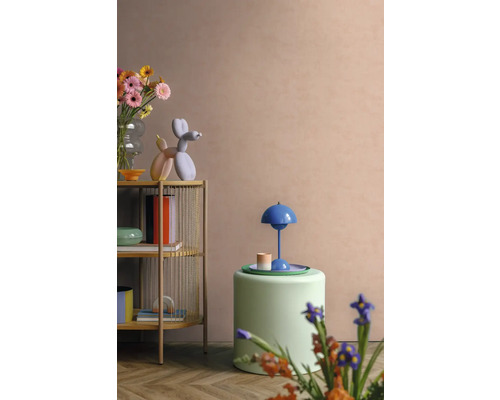 Modernes Wohnzimmer mit hellem Holzregal, farbenfrohen Dekorationsobjekten, grünem Polsterhocker und einer blauen Tischleuchte vor einer pfirsichfarbenen Wand.