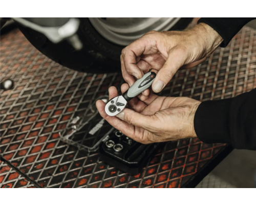 Hände halten eine WGB Bit-Ratsche über einer Werkbank in einer Werkstatt beim Schrauben an einem Motorrad.