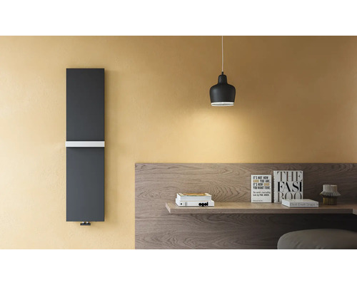 Moderner vertikaler Designheizkörper in Dunkelgrau mit weißem Handtuchhalter an einer gelben Wand neben einem Holztisch.