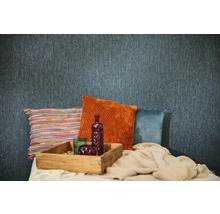 Strukturierte blaue Tapete in einer Raumszene mit dekorativen Kissen in Orange, Blau und Bunt, einem Holztablett und einer gestrickten Decke.