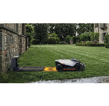 Mähroboter Mammotion auf einem grünen Rasen nähert sich der Ladestation bei Regen vor einer Hauswand aus Naturstein.