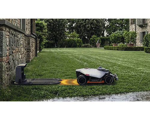 Mähroboter Mammotion auf einem grünen Rasen nähert sich der Ladestation bei Regen vor einer Hauswand aus Naturstein.