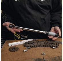 Drehmomentschlüssel aus Metall mit verstellbarem Griff, präsentiert von einer Person über einer Werkbank mit Kette und Werkzeugen.