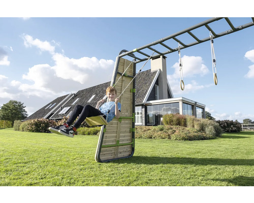 Kind auf einer Schaukel an einem Klettergerüst mit Hangelstangen und Holzwand in einem hellen, geräumigen Garten vor einem Wohnhaus.