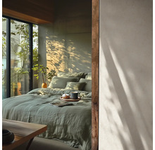 Helles Schlafzimmer mit grüner Leinenbettwäsche auf dem Bett, großen Fenstern und Schatten von Pflanzen an der Wand. Minimalistischer Stil.