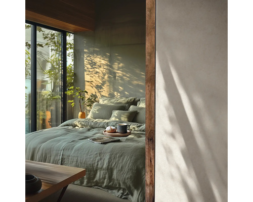 Helles Schlafzimmer mit grüner Leinenbettwäsche auf dem Bett, großen Fenstern und Schatten von Pflanzen an der Wand. Minimalistischer Stil.