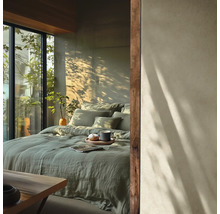 Helles Schlafzimmer mit salbeigrüner Leinenbettwäsche auf einem großen Bett vor einer Betonwand mit Sonnenlicht und Blick ins Grüne.