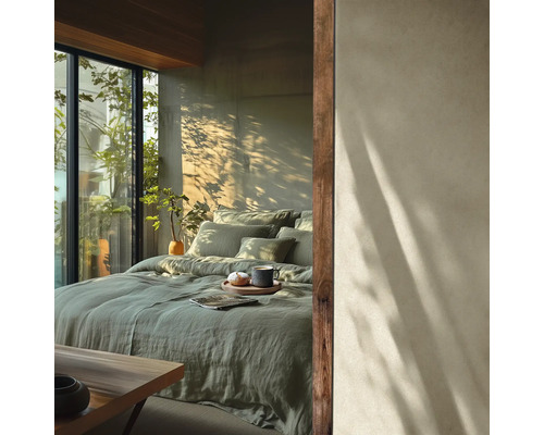 Helles Schlafzimmer mit salbeigrüner Leinenbettwäsche auf einem großen Bett vor einer Betonwand mit Sonnenlicht und Blick ins Grüne.