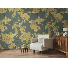 Vlies-Fototapete mit gelben Blütenzweigen auf dunklem Hintergrund im Wohnzimmer mit hellem Sessel und Holzmöbeln auf Laminatboden.