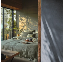 Helles Schlafzimmer mit grünem Leinenbettzeug auf dem Bett, strukturierter Wand und Holzakzenten neben einem Fenster mit Blick ins Grüne.