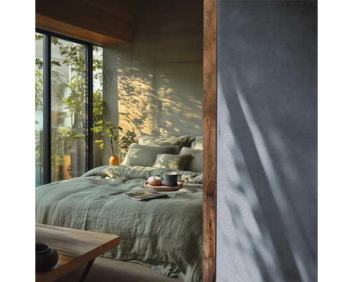 Helles Schlafzimmer mit grünem Leinenbettzeug auf dem Bett, strukturierter Wand und Holzakzenten neben einem Fenster mit Blick ins Grüne.
